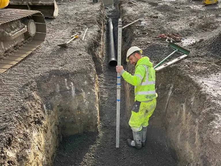 Worker measuring depth in a drainage trench next to an excavator
