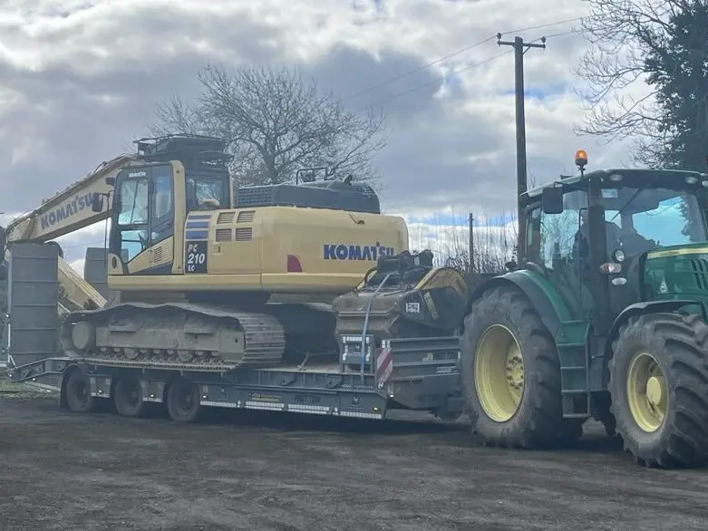 Komatsu PC210 excavator loaded on a trailer hitched to a John Deere tractor